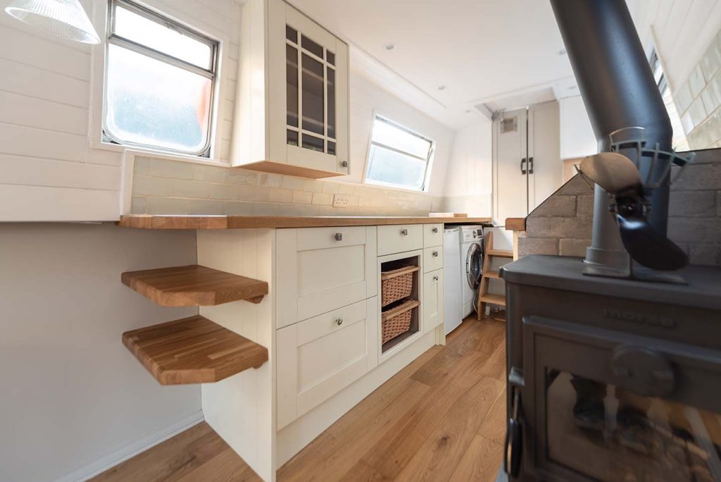 Narrowboat galley style kitchen.  White shaker cabinets with oak butchers block worktop and shelves. Glass fronted cabinet on wall above. Multi fuel stove in the fore-ground