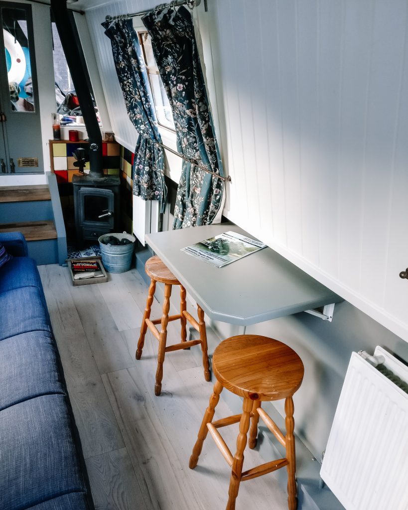 Narrowboat salon, showing fold up table wth stools and sofa.  Light grey beneath the gunwale and qhit above. Multi fuel stove in the corner next to storage steps
